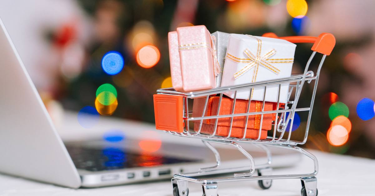 Miniatureinkaufswagen mit Miniaturgeschenken gef&uuml;llt neben einem aufgeklappten Laptop. Im Hintergrund ein geschm&uuml;ckter Weihnachtsbaum